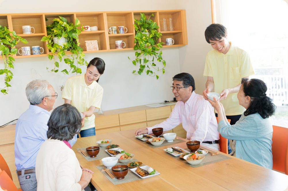 介護施設内で昼食を楽しむ4人の利用者と2人の介護職員